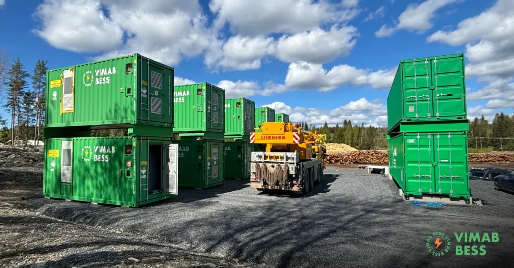 Gröna förvaringsbehållare märkta VIMAB BESS, en del av ett internationellt batterilagringsavtal, är staplade utomhus på grus. En gul Liebherr-kranbil står mellan dem, med träd och en molnig himmel i bakgrunden. VIMAB BESS-logotypen syns längst ner till höger.